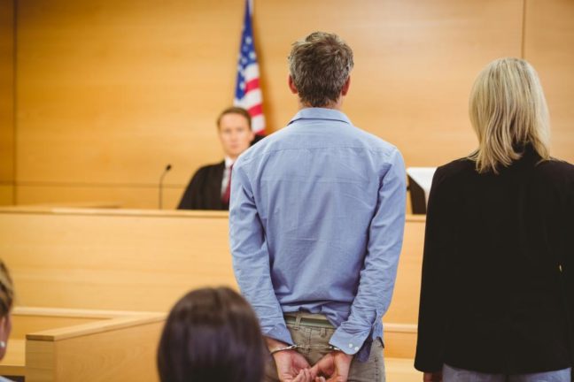 Man in handcuffs standing before a judge in a courtroom, with a lawyer beside him, discussing criminal defense matters.