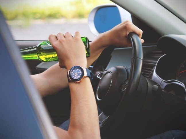 Person holding a green beer bottle while driving, illustrating the dangers of DUI.
