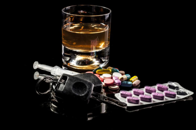Glass of whiskey, assorted pills, car keys, and a syringe, representing DUI implications and substance influence on driving.