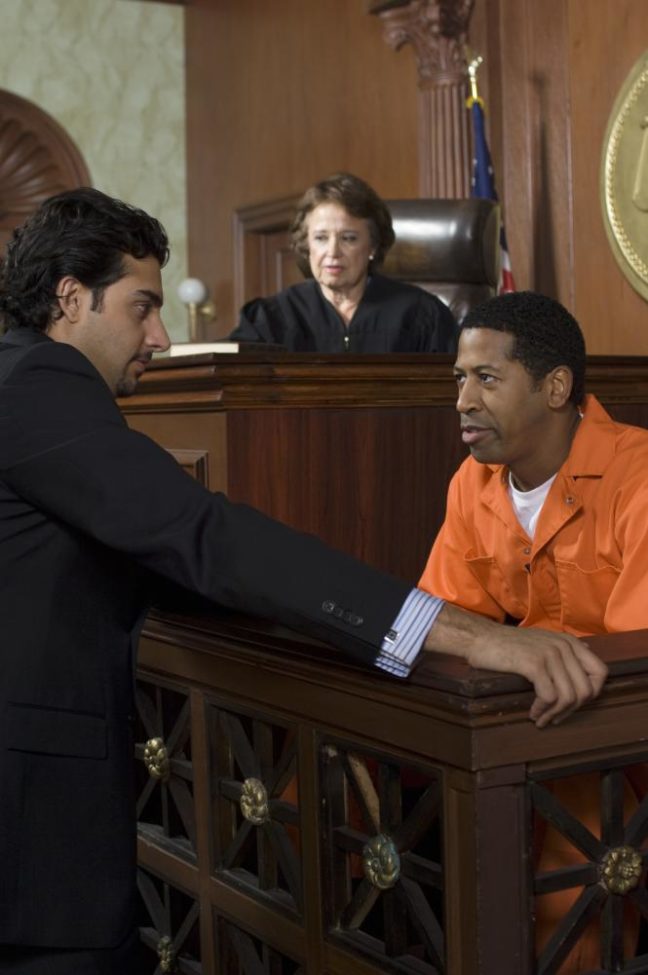 Judge overseeing a court case with a lawyer consulting a defendant in an orange jumpsuit, emphasizing legal representation in criminal defense matters.