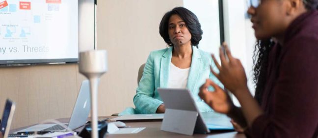 Professional woman in a light blue blazer listening attentively during a discussion in a conference room, with a presentation on the screen about opportunities and threats.