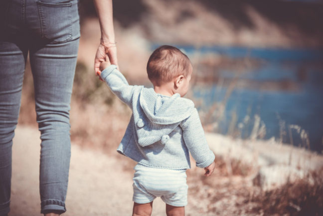 Toddler in a blue sweater and white shorts holding an adult's hand, walking along a scenic path by the water, symbolizing family connections and the journey of adoption.