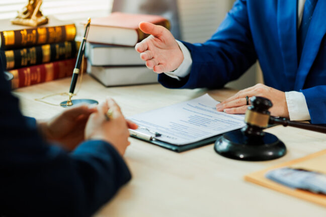 Lawyer in blue suit discussing legal documents with client, gavel and law books on table, emphasizing trial experience and legal advocacy.